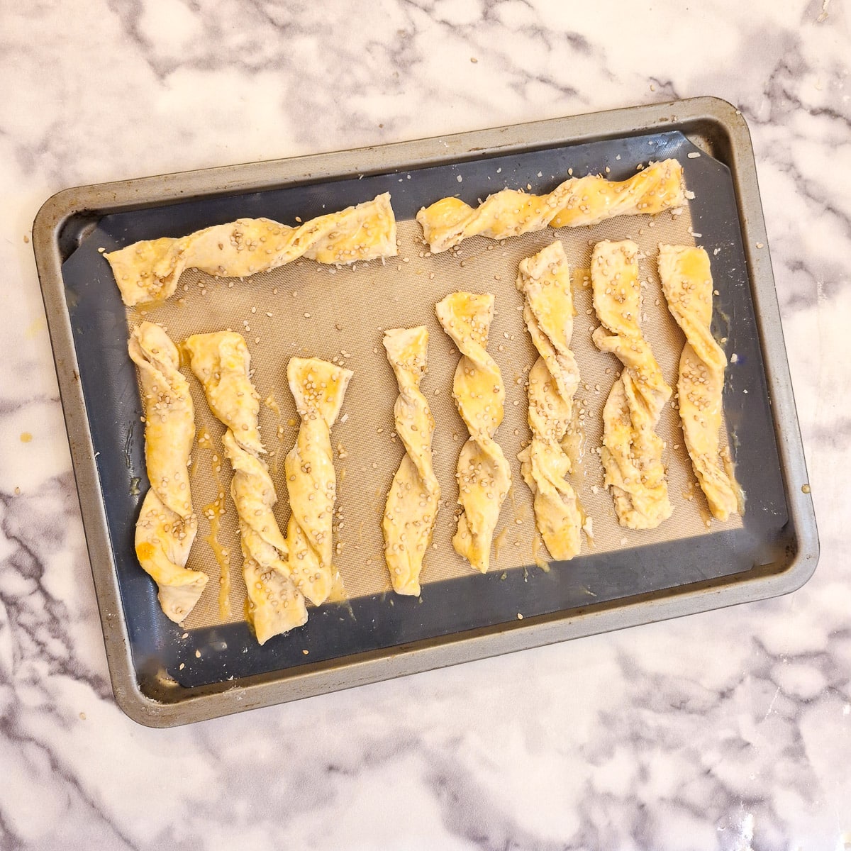 Unbaked cheese straws brushed with beaten egg and sprinkled with sesame seeds, on a baking sheet.