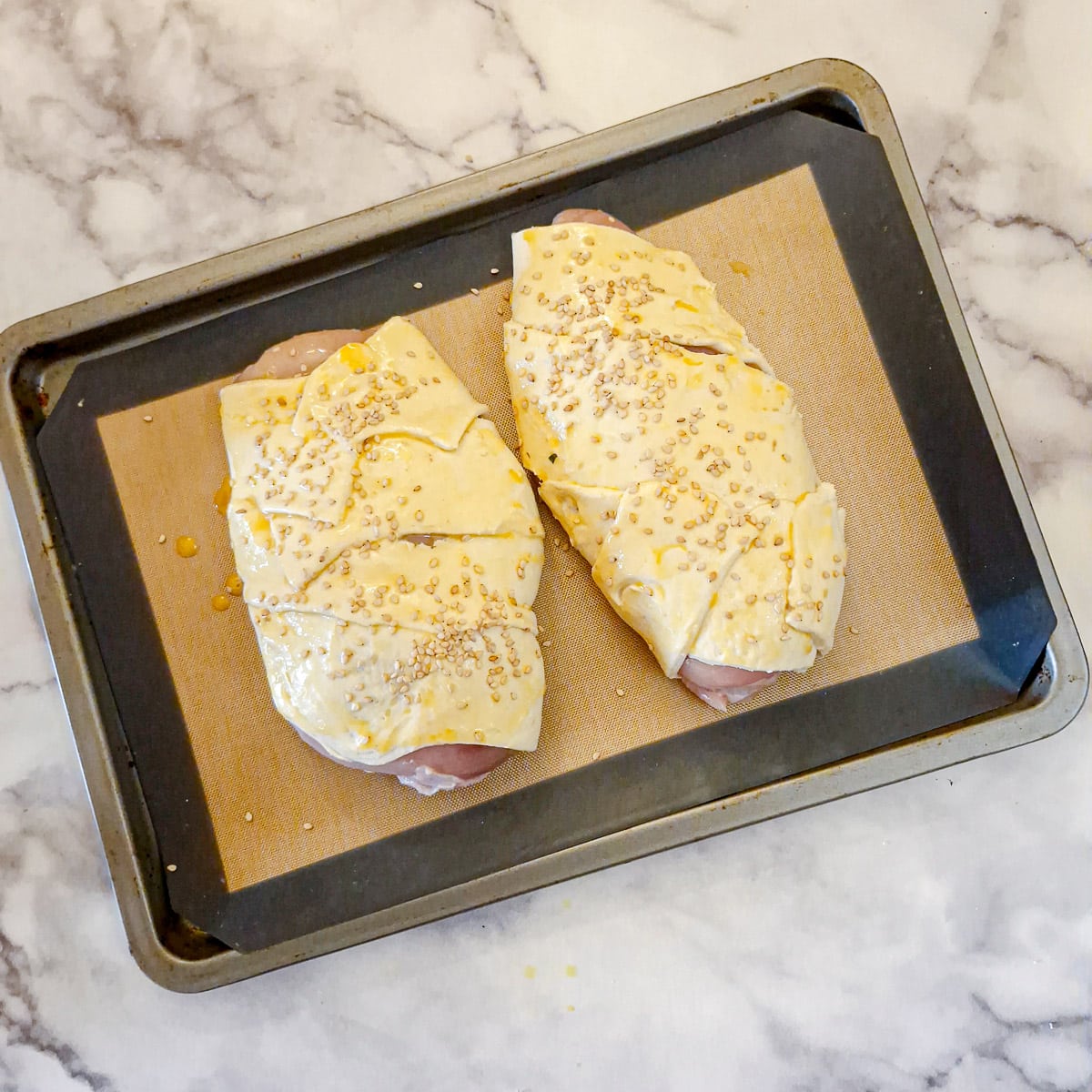 Mushroom-stuffed chicken en croute brushed with egg-wash and sprinkled with sesame seeds on a baking tray.