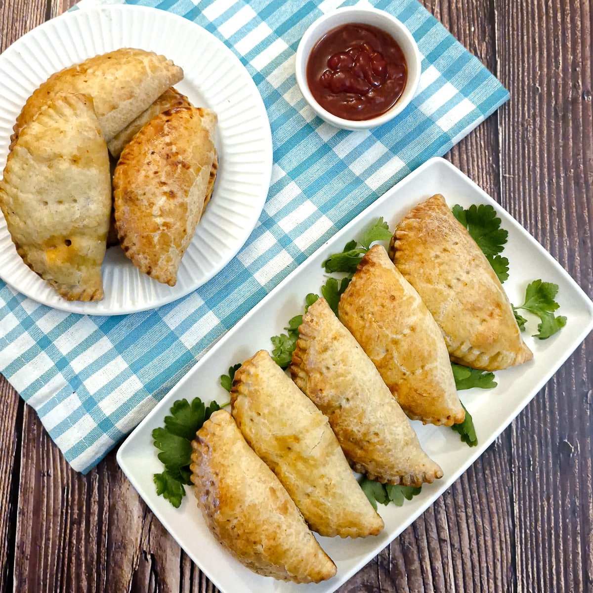 5 empanadas on a white oblong plate with more empanadas and a dish of ketchup in the background.