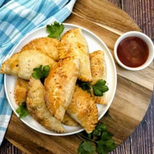 A pile of chicken empanadas on a round white plate alongside a small dish of dipping sauce.