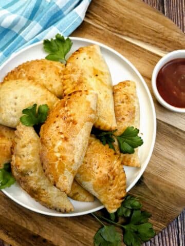 A pile of chicken empanadas on a round white plate alongside a small dish of dipping sauce.