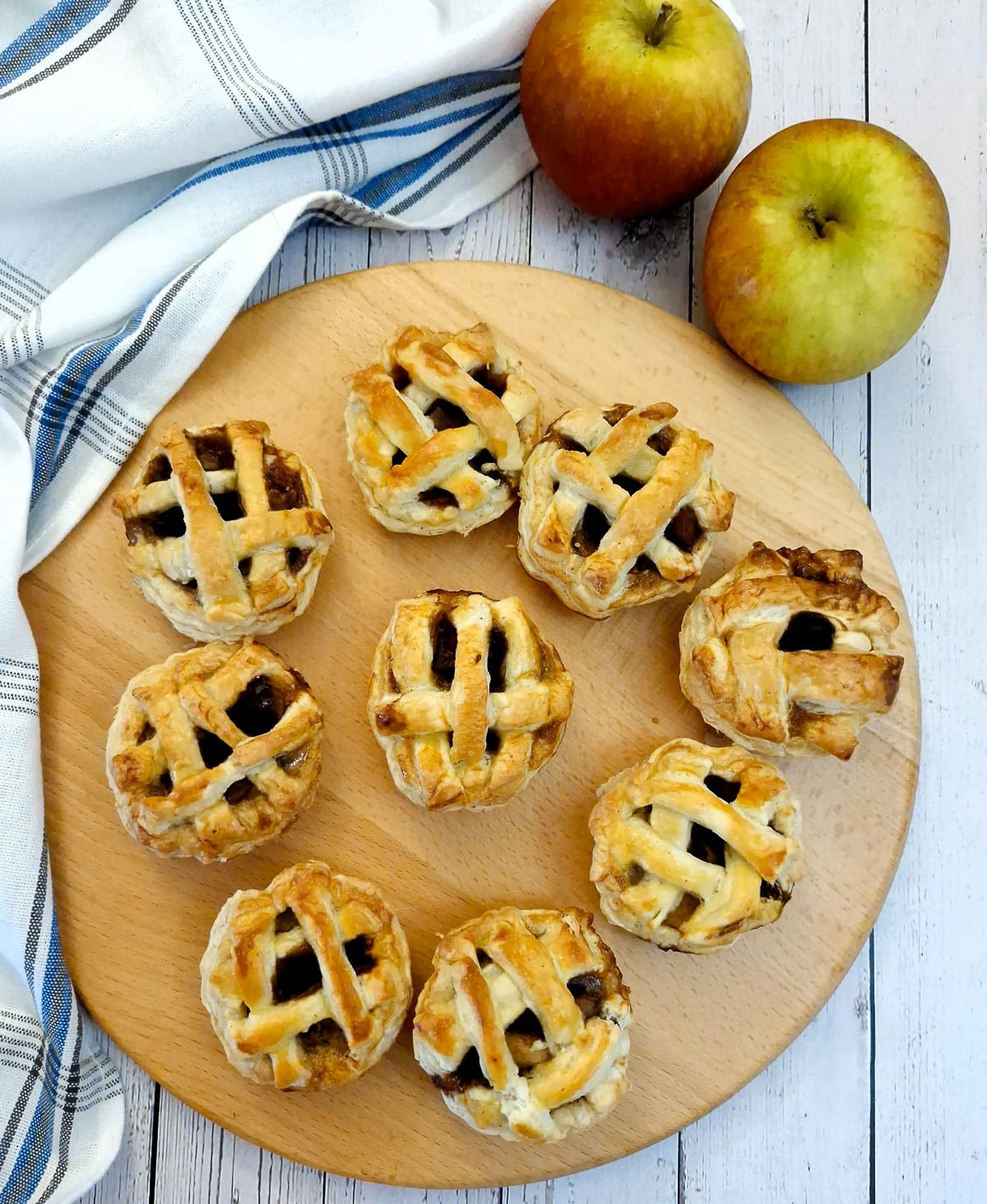 9 caramel apple pies on a wooden board with 2 apples in the background.
