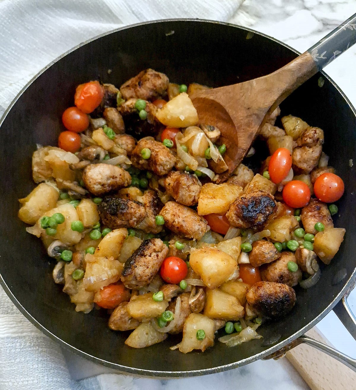 A frying pan of potato and sausage hash with a large wooden serving spoon.