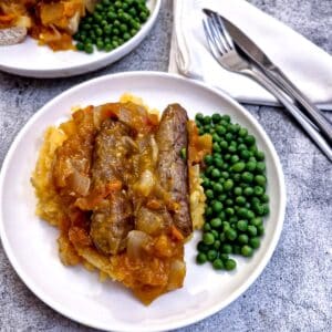 A plate of sausages on a bed of sweet potato mash covered in tomato and onion gravy with a side helping of peas.
