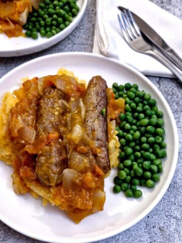 A plate of sausages on a bed of sweet potato mash covered in tomato and onion gravy with a side helping of peas.