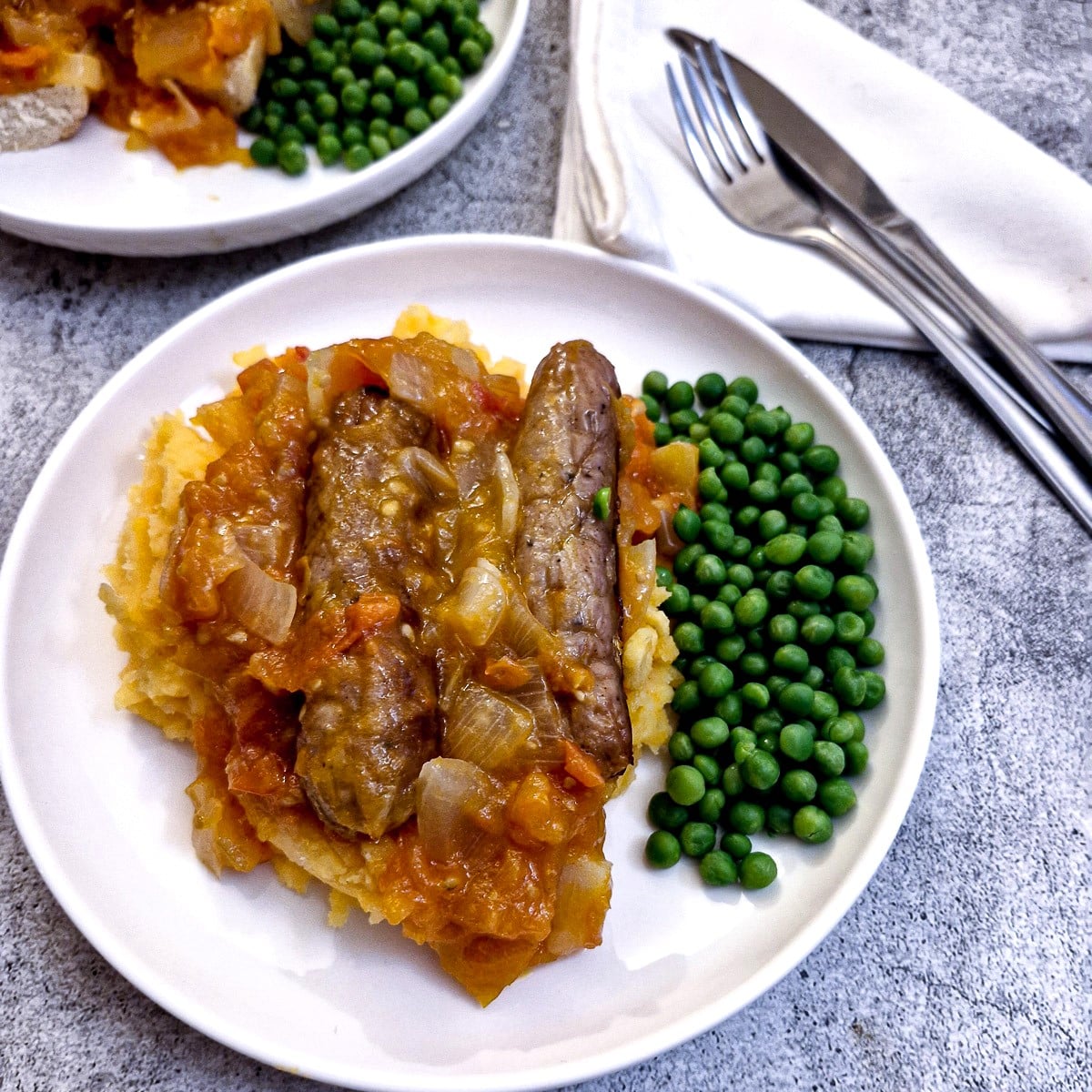 A plate of sausages on a bed of sweet potato mash covered in tomato and onion gravy with a side helping of peas.