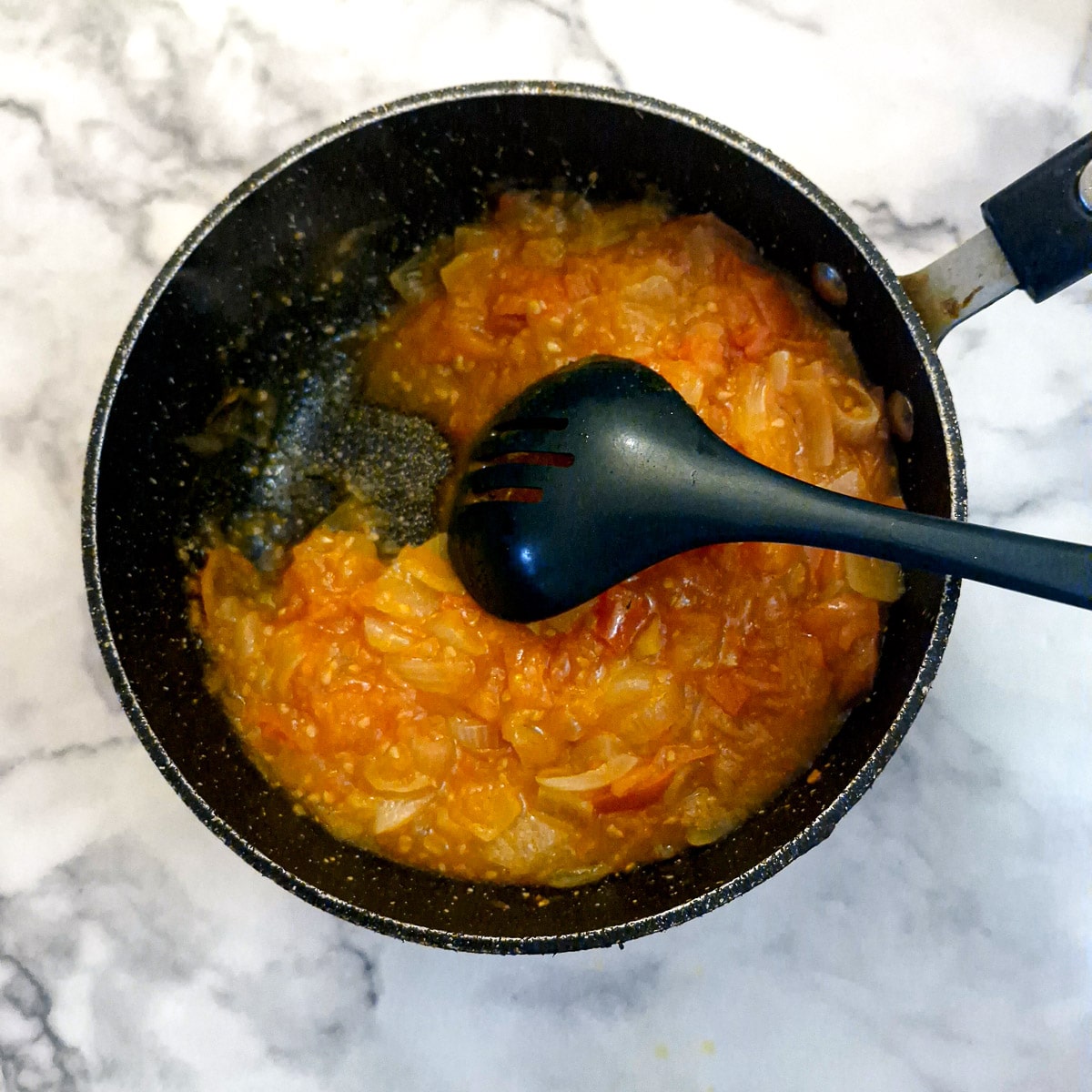 A spoon being drawn through a pan of tomato and onion gravy to show the thick consistency.