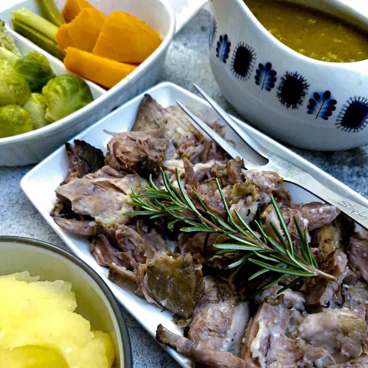 A plate of shredded lamb shoulder with dishes of vegetables and gravy.