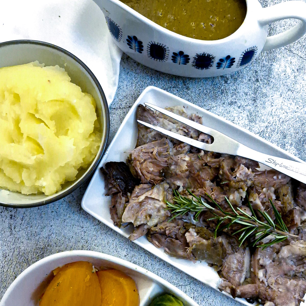 Lamb shoulder on a serving plate surrounded by dishes of vegetables and gravy.
