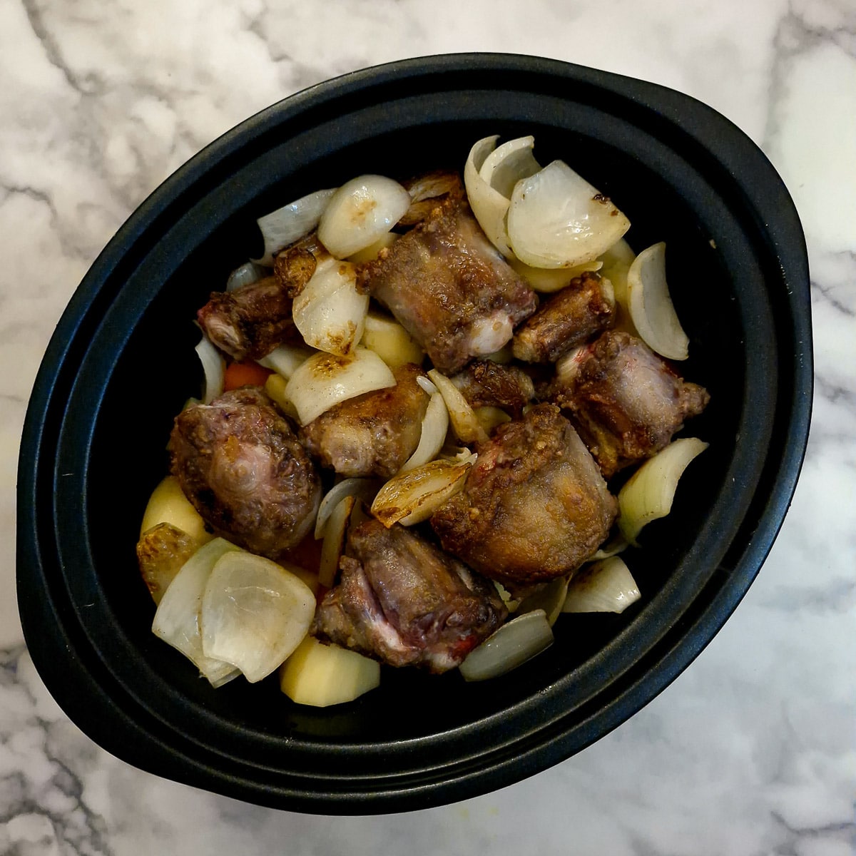 Oxtail and onions arranged on top of vegetables in a slow cooker.