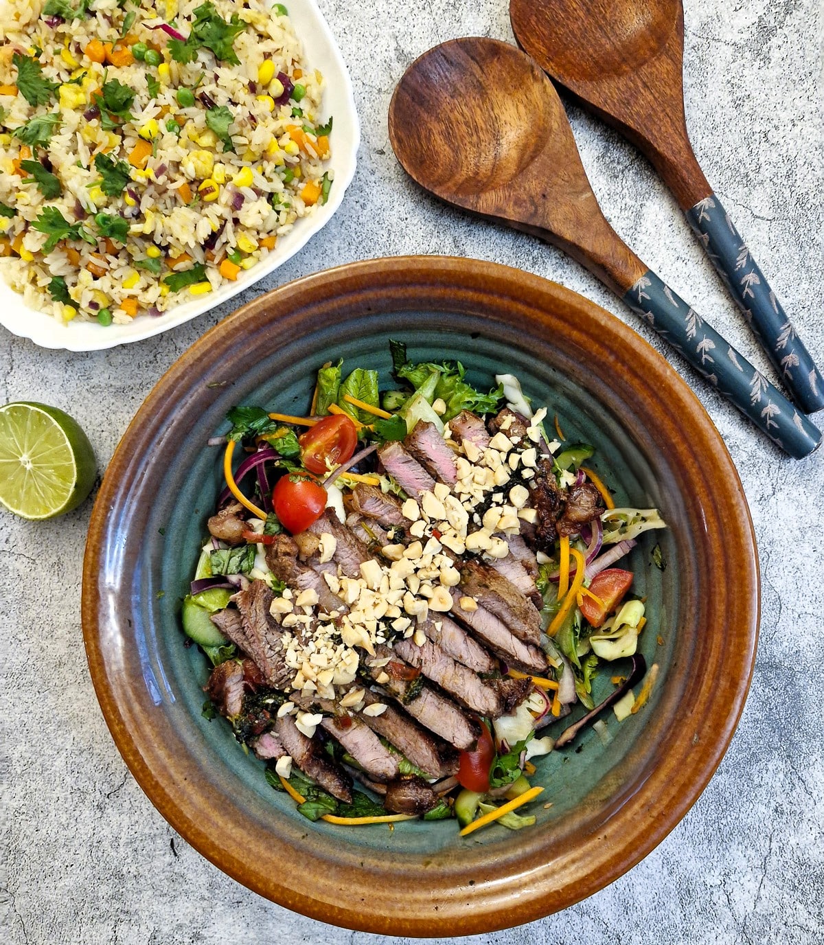 A serving dish of Thai beef salad with a dish of rice in the background next to 2 wooden serving spoons.