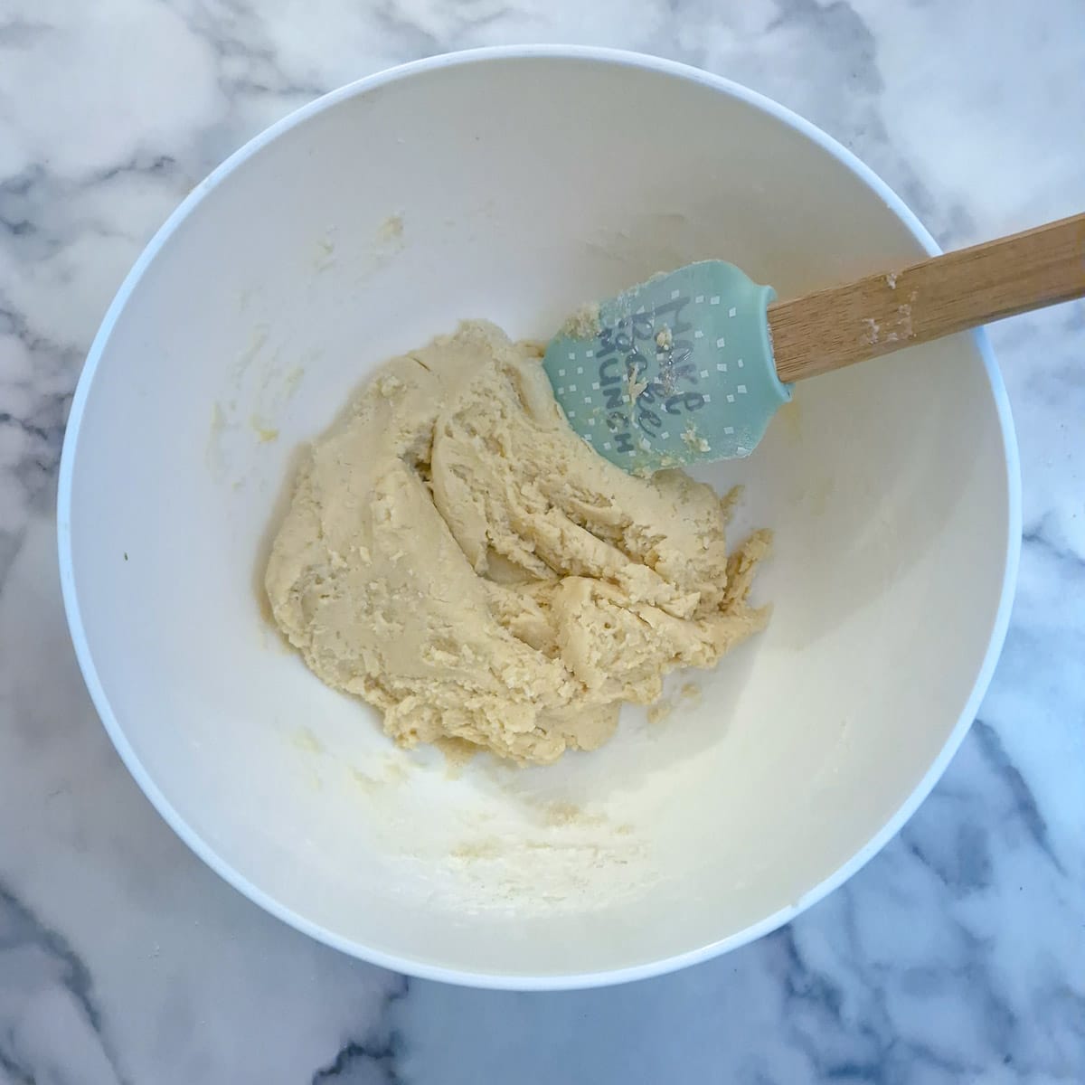 Biscotti dough being mixed in a white mixing bowl with a spatula.