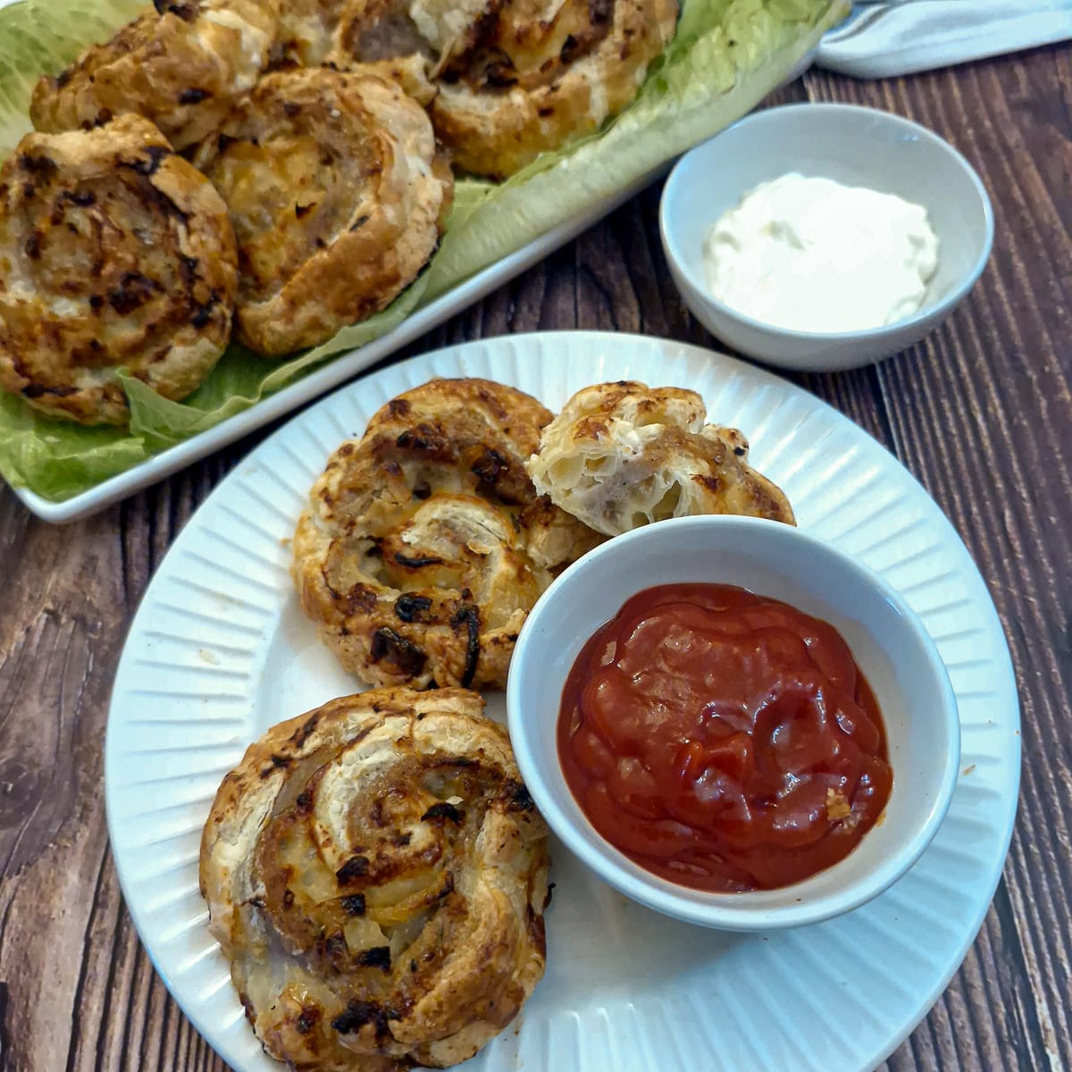 Sausage and onion pinwheels on a white plate with a small dish  of tomato ketchup for dipping.  There is a plate of sausage pinwheels in the background.