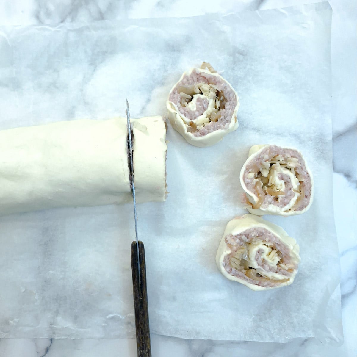 Pinwheels being cut from a rolled puff pastry sausage roll.