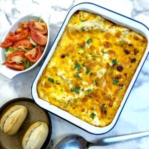 A white enamel baking dish filled with cheese-toppped lasagne. There is a dish of tomato salad in the background and two crusty rolls on a plate.