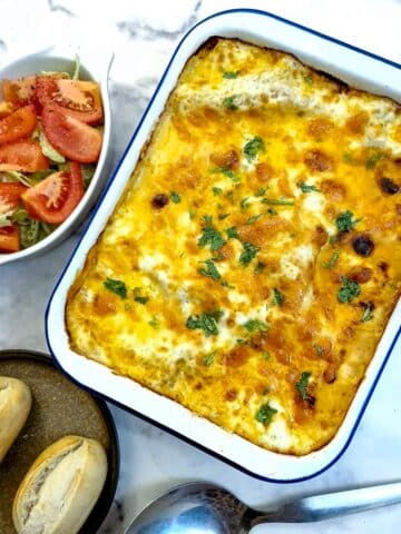 A white enamel baking dish filled with cheese-toppped lasagne. There is a dish of tomato salad in the background and two crusty rolls on a plate.