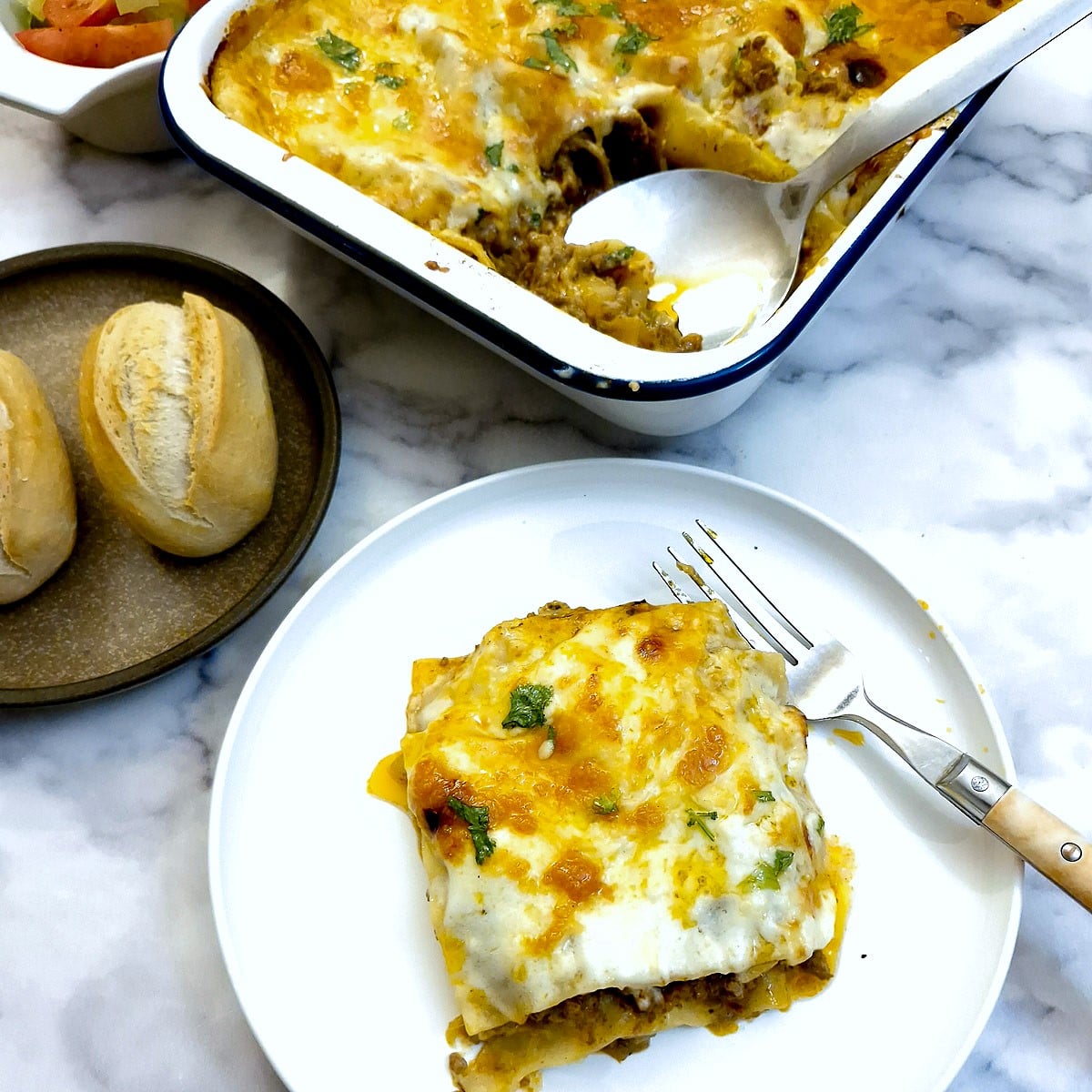 A slice of lasagne on a white plate with a fork.  There is a dish of lasagne in the background.