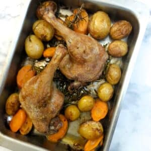 2 oven-roasted duck legs in a baking dish with potatoes and carrots.