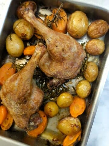 2 oven-roasted duck legs in a baking dish with potatoes and carrots.