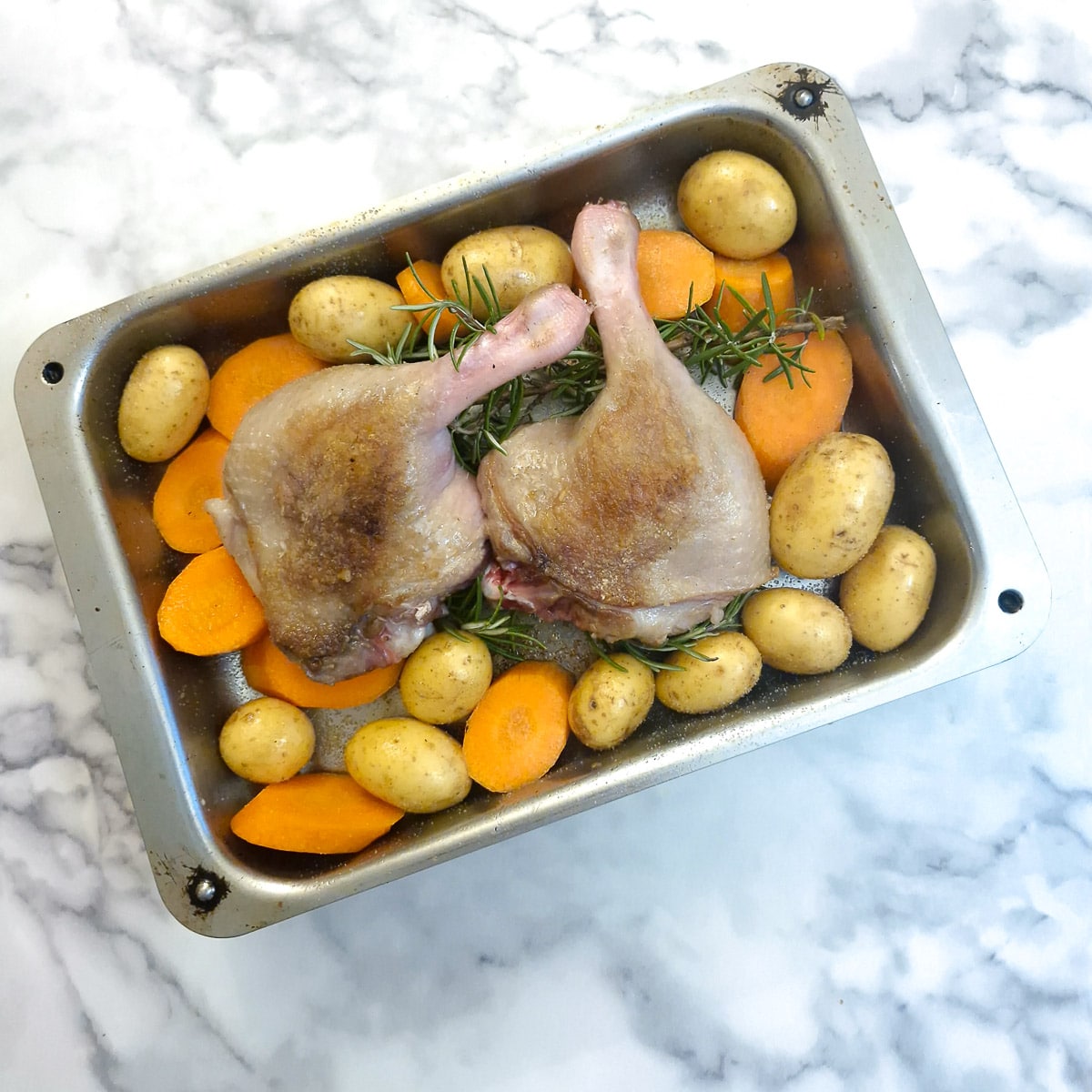 2 duck legs on bed of rosemary surrounded by potatoes and carrots in a roasting pan.