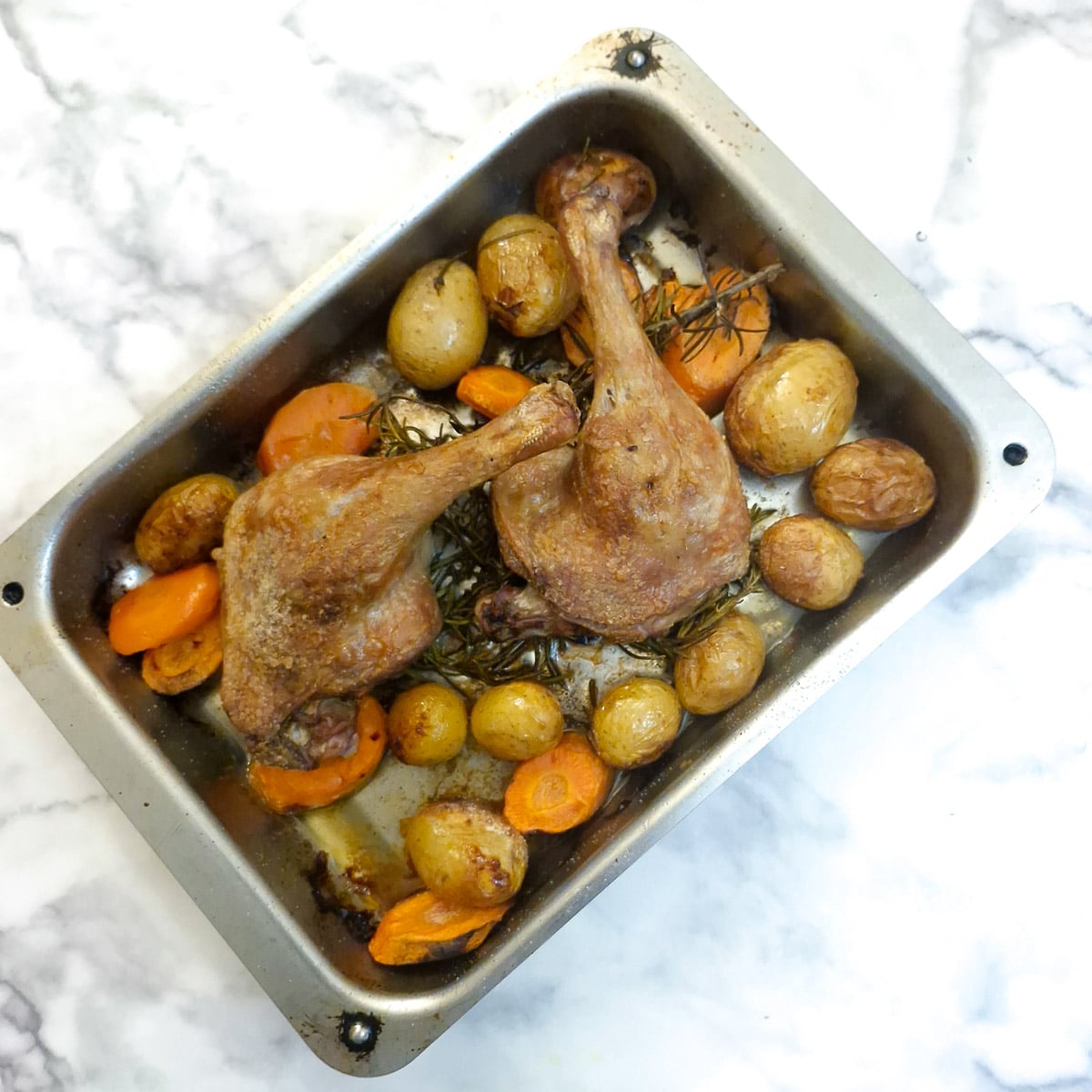Two cooked duck legs with potatoes and carrots in a roasting pan.