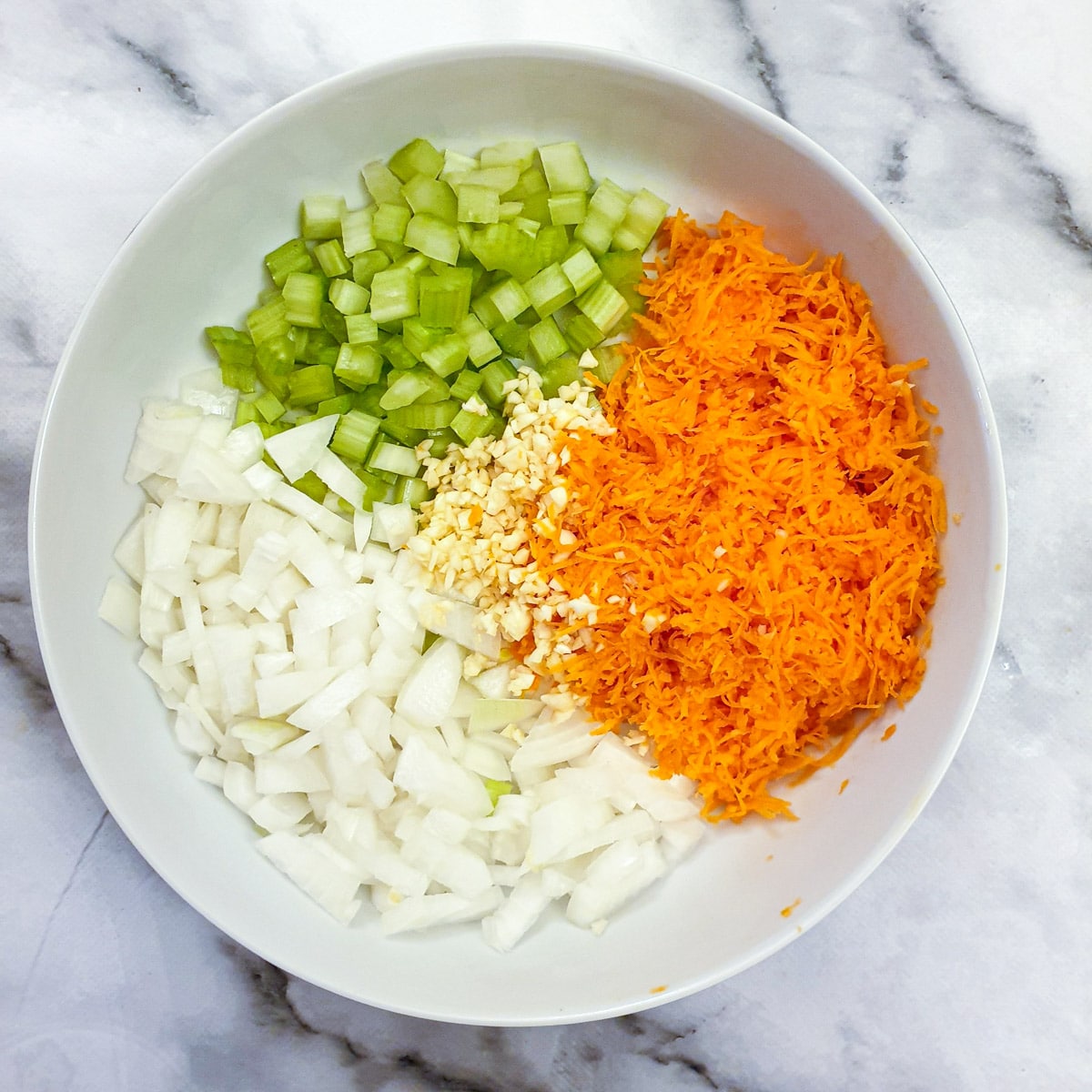 Chopped onions and celery with grated carrots and minced garlic in a white dish.