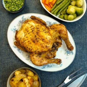 An air fryer roast chicken on a serving plate surrounded by dishes of vegetables.