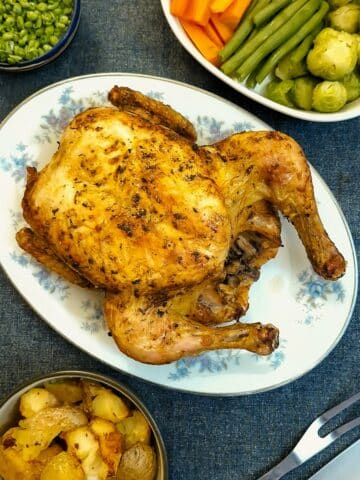 An air fryer roast chicken on a serving plate surrounded by dishes of vegetables.