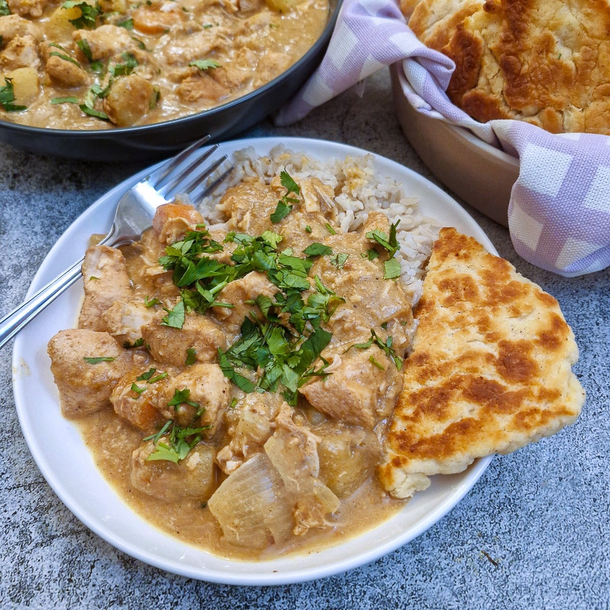 A white dinner plate with brown rice topped with chicken curry.  There is a piece of naan bread on the side.