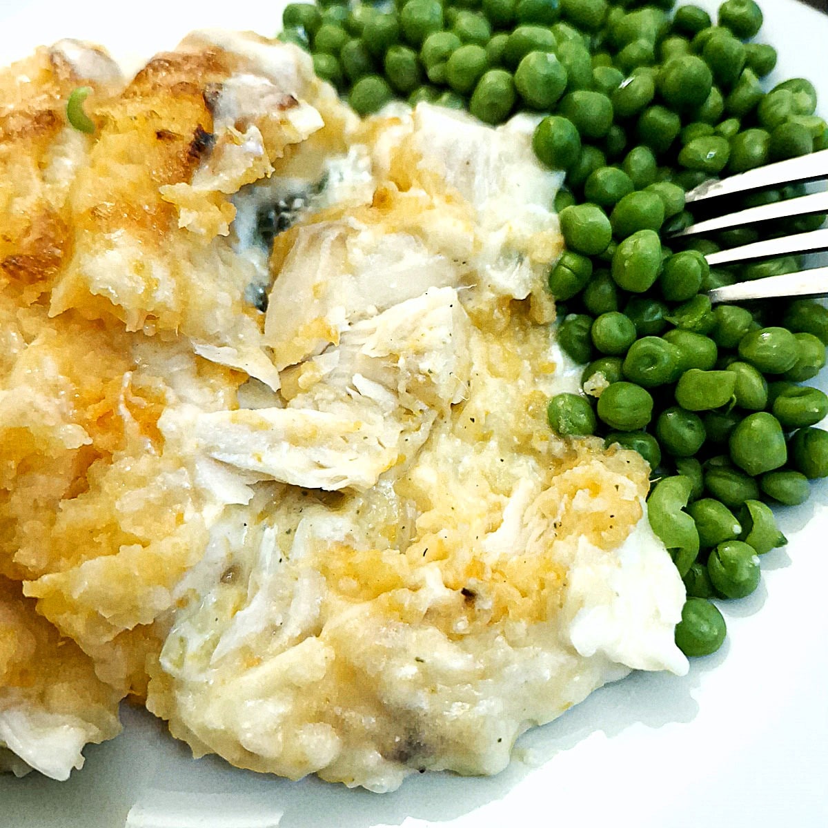 Close up of haddock au gratin on a white place alongside a serving of peas.