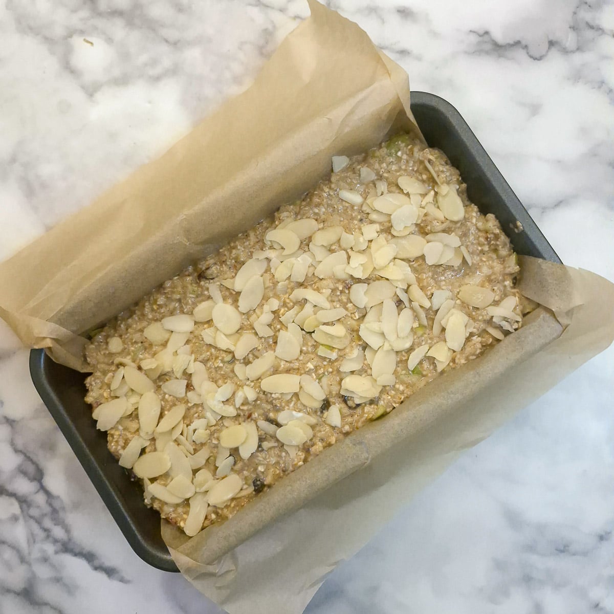 An unbaked honey oat apple and banana loaf topped with flaked almonds in a baking dish which has been lined with baking paper.