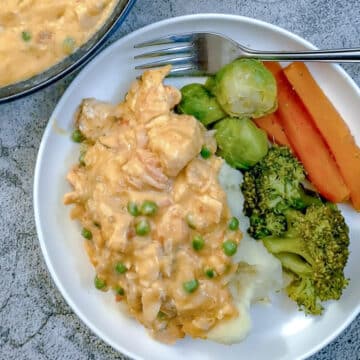 A white dinner plate with mashed potatoes covered in poached salmon in sauce with carrots, broccoli and brussels sprouts on the side.
