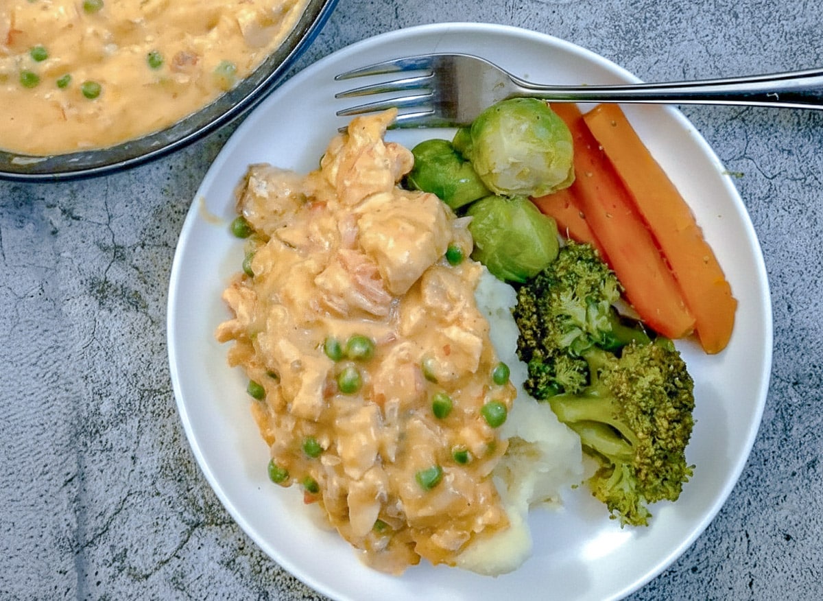 A white dinner plate with mashed potatoes covered in poached salmon in sauce with carrots, broccoli and brussels sprouts on the side.