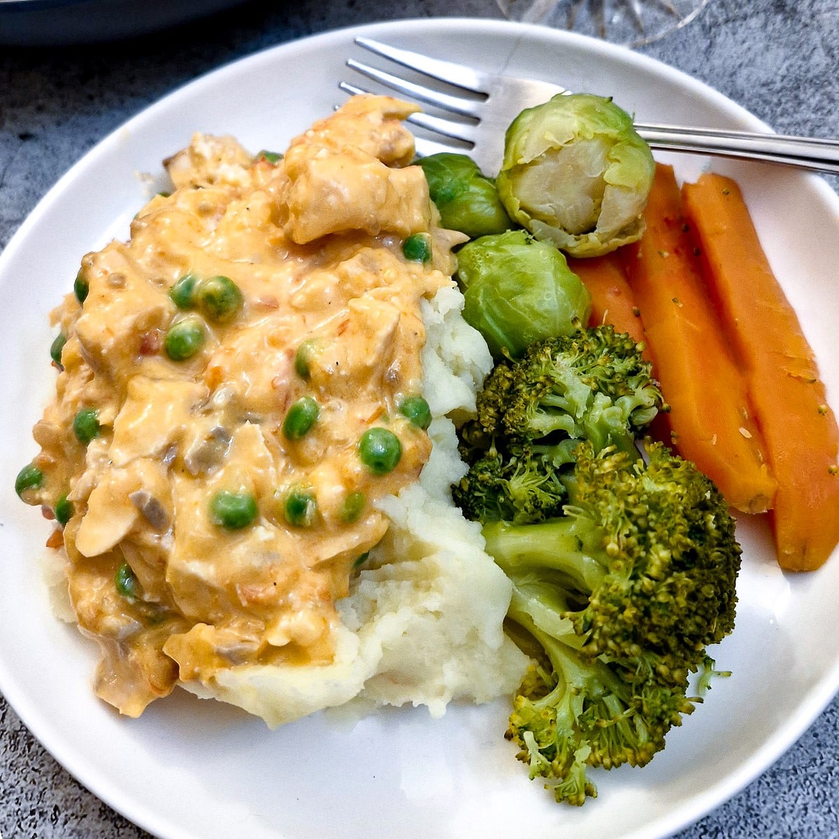 A white dinner plate with mashed potatoes covered in poached salmon in sauce with carrots, broccoli and brussels sprouts on the side.