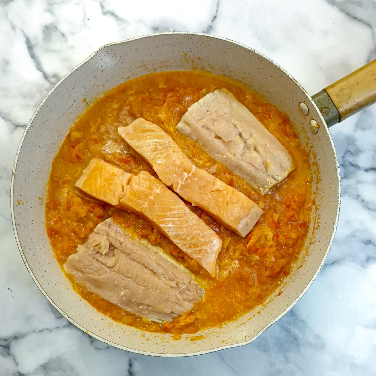 Raw salmon fillets nestled in red pepper sauce in a frying pan.