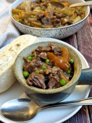 A dish of beef stew in a soup bowl on a white plate with a crusty breadroll. There is a dish of stew in the background.