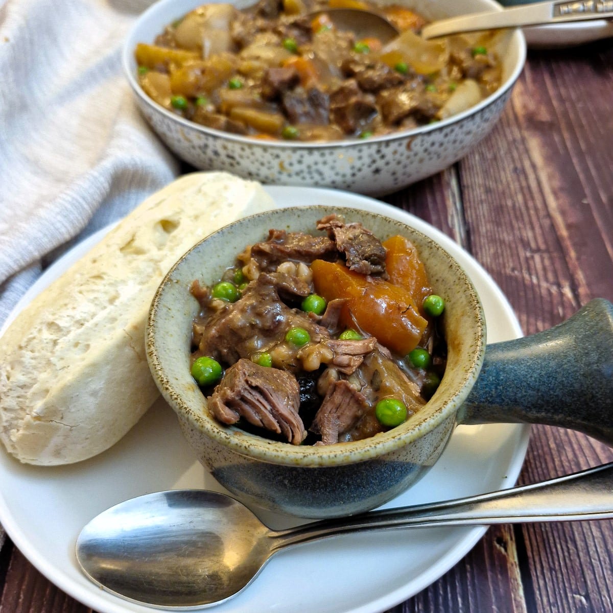 A dish of beef stew in a soup bowl on a white plate with a crusty breadroll. There is a dish of stew in the background.