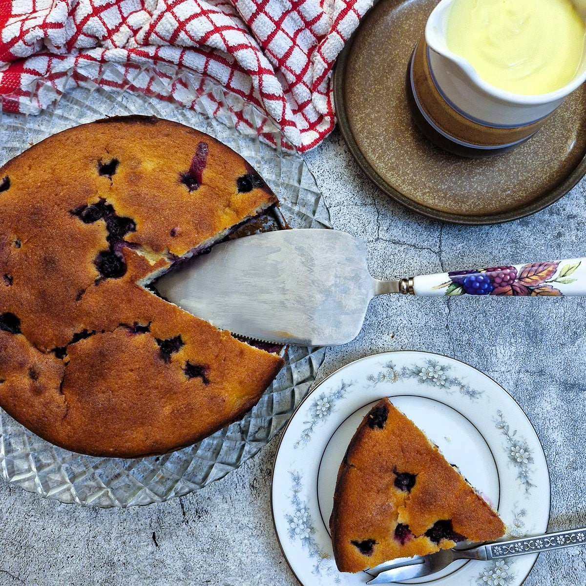 Overhead shot of a blueberry sponge cake alongside a jug of custard.  There is a slice of cake on a plate in the foreground.