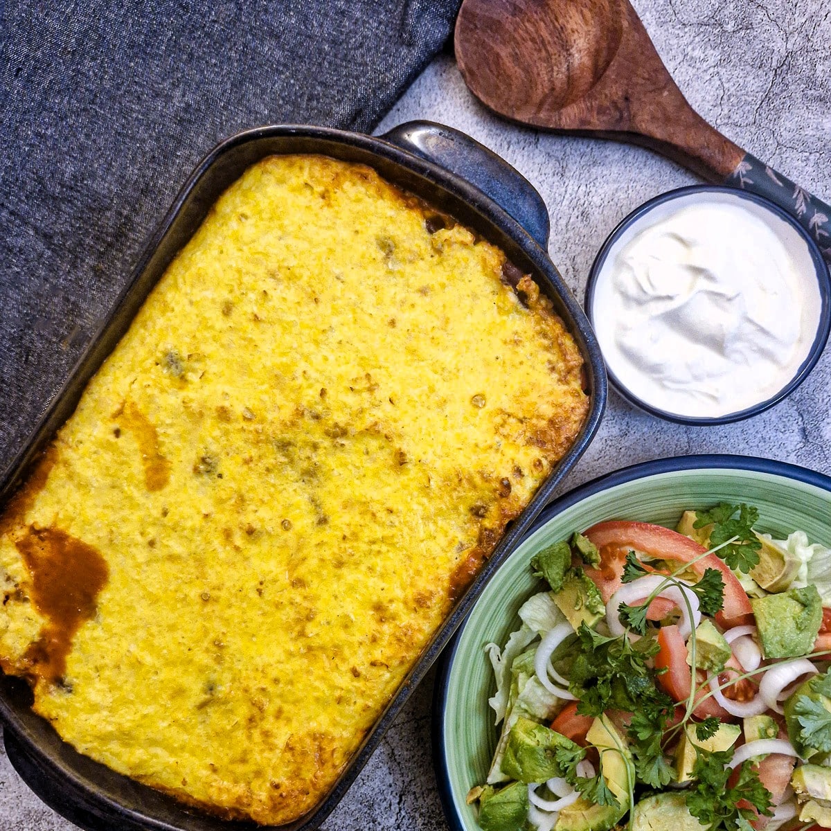 Overhead shot of a dish of chilli con carne rice casserole with dishes of salad and sour cream on the side.