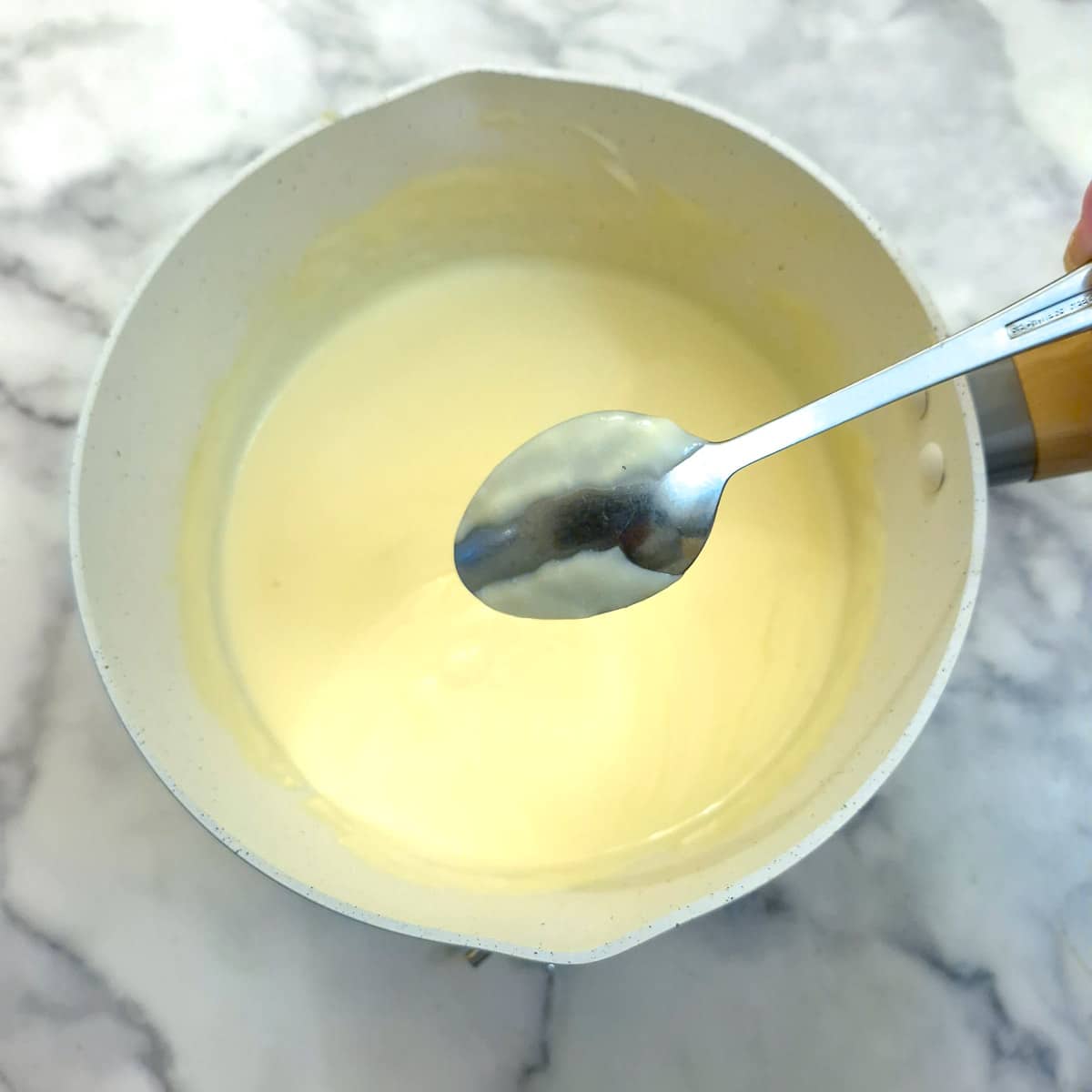 A spoon held over a pan of vanilla custard showing how the custard coats the spoon.