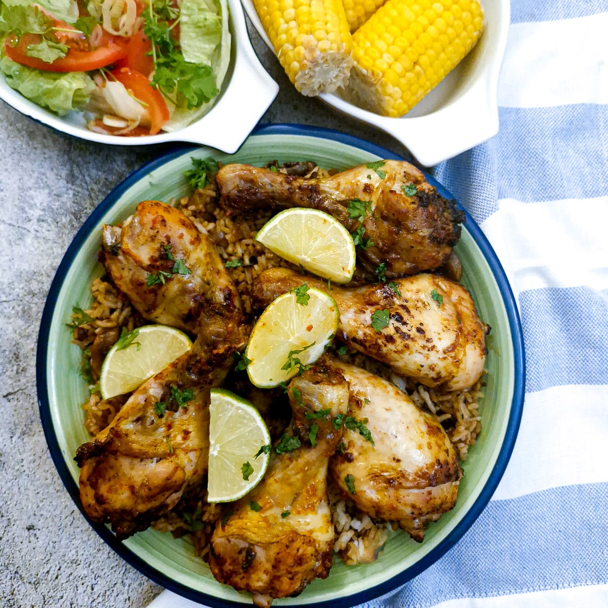 Overhead shot of a dish of peri peri chicken drumsticks with a dish of corn and a green salad in the background.