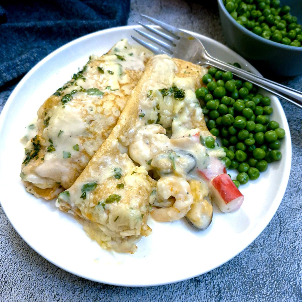 2 seafood stuffed pancakes with peas on a white plate.  One pancake has been cut in half to show the filling.