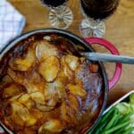 Steak and kidney hotpot in a red casserole dish. There are 2 glasses of red wine in the background.