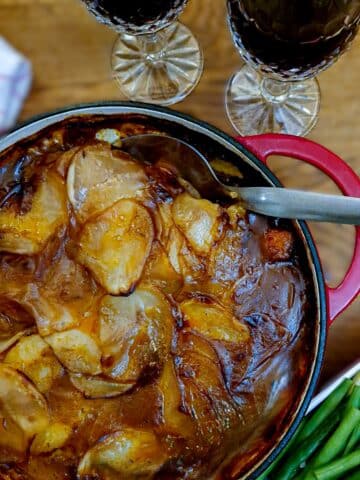 Steak and kidney hotpot in a red casserole dish. There are 2 glasses of red wine in the background.