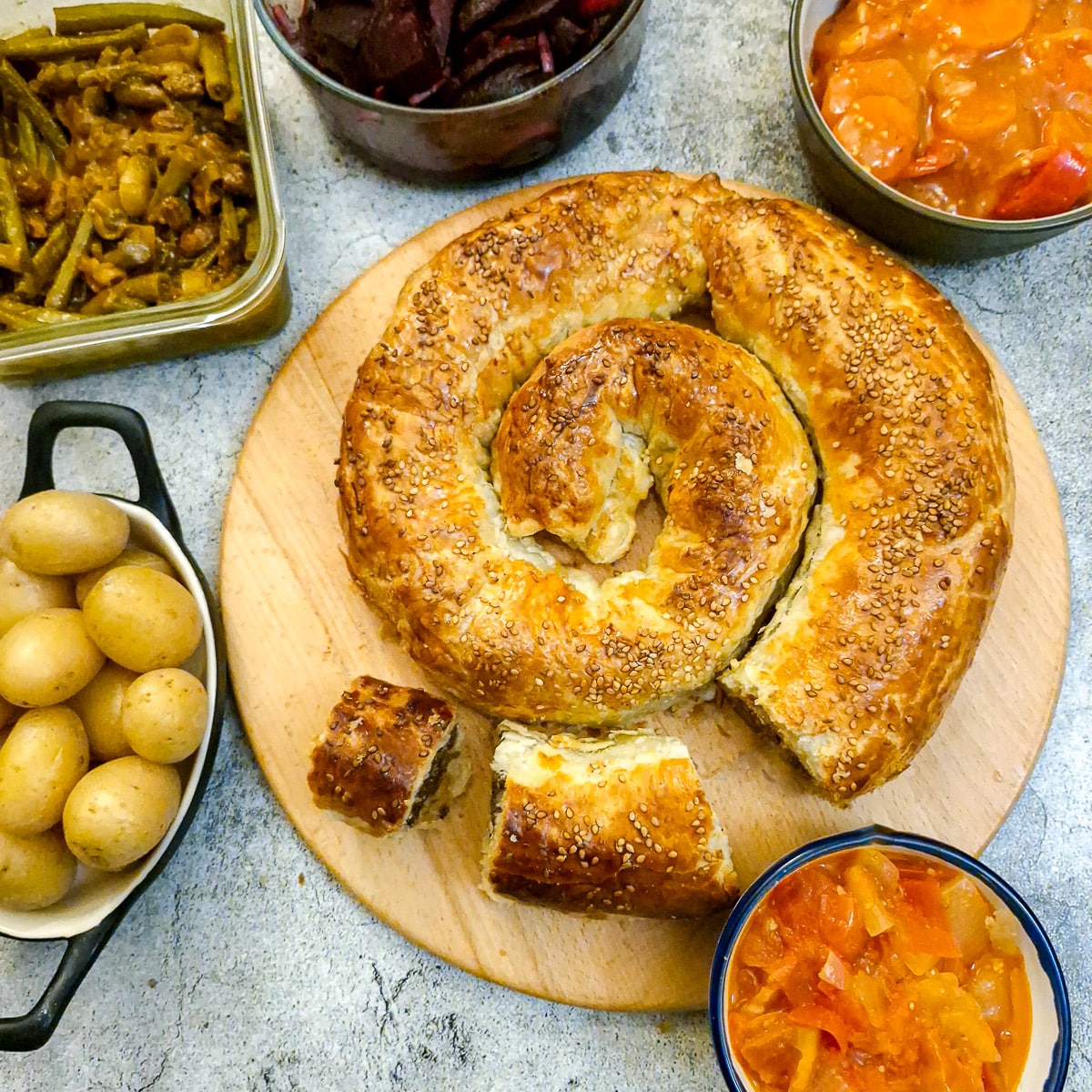 A boerewors puff pastry spiral on a wooden board surrounded by a selection of salads.