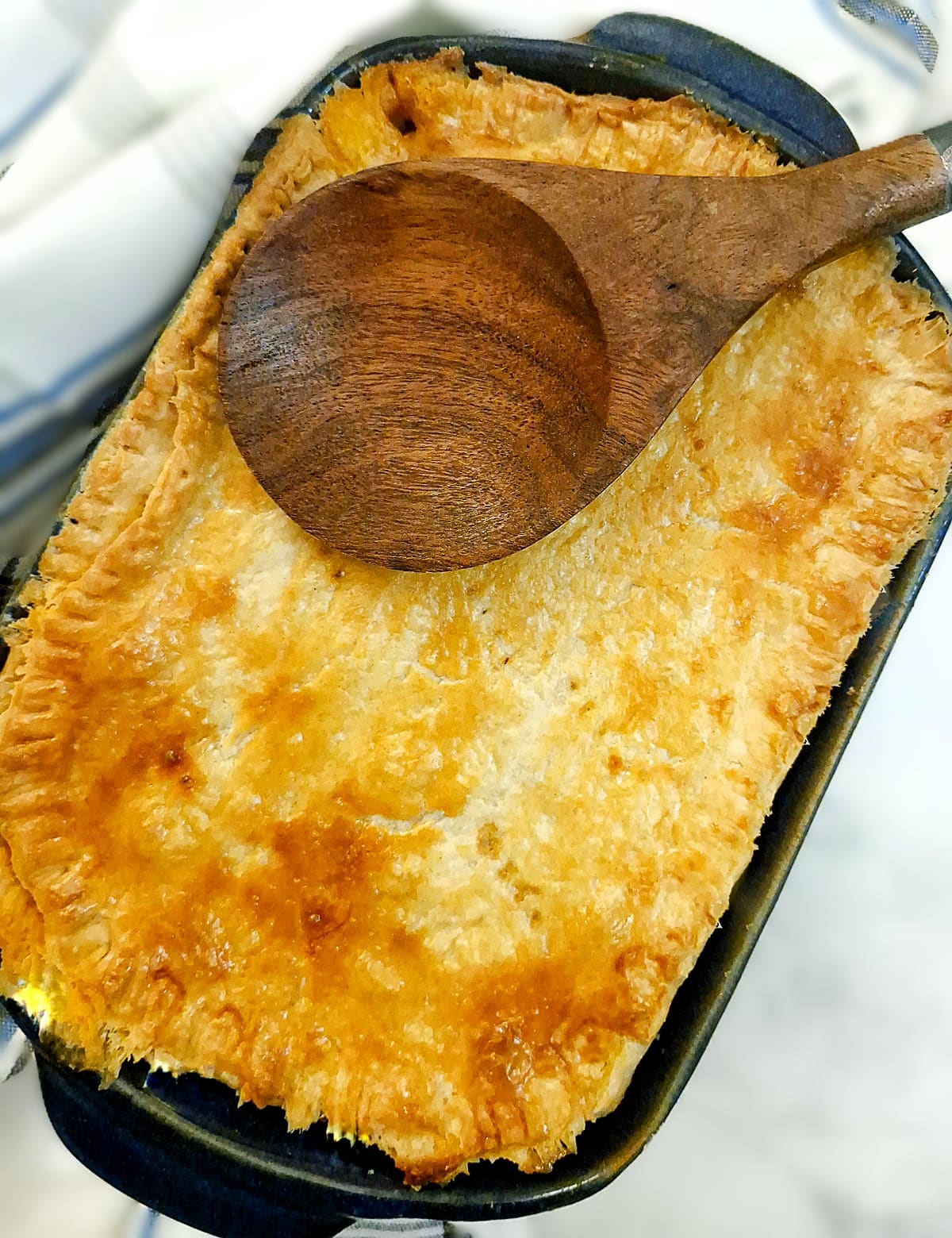 A browned meat pie in a baking dish with  wooden serving spoon on top.