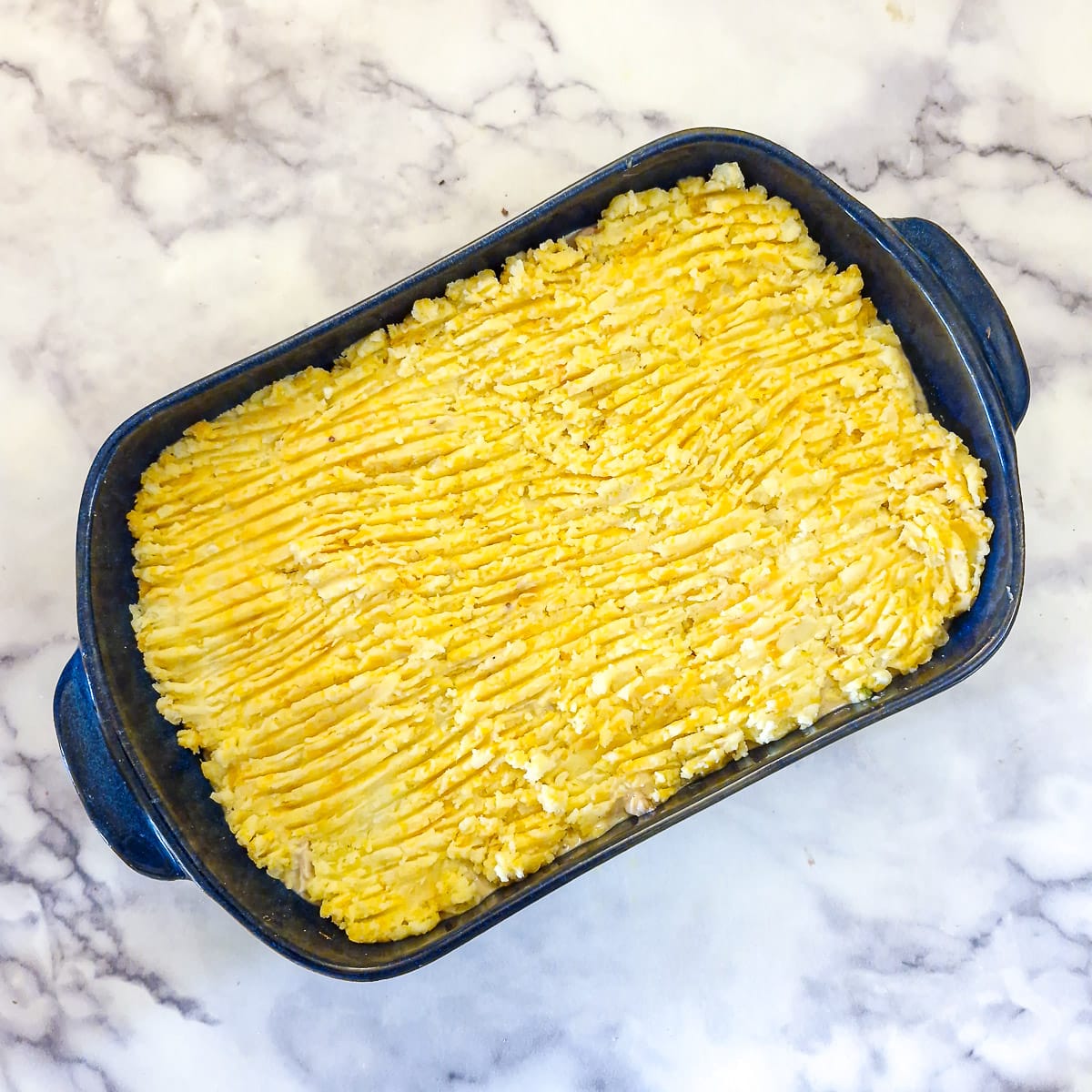 Potato mash that has been smoothed out with a fork on top of the chicken filling in a blue baking dsh.