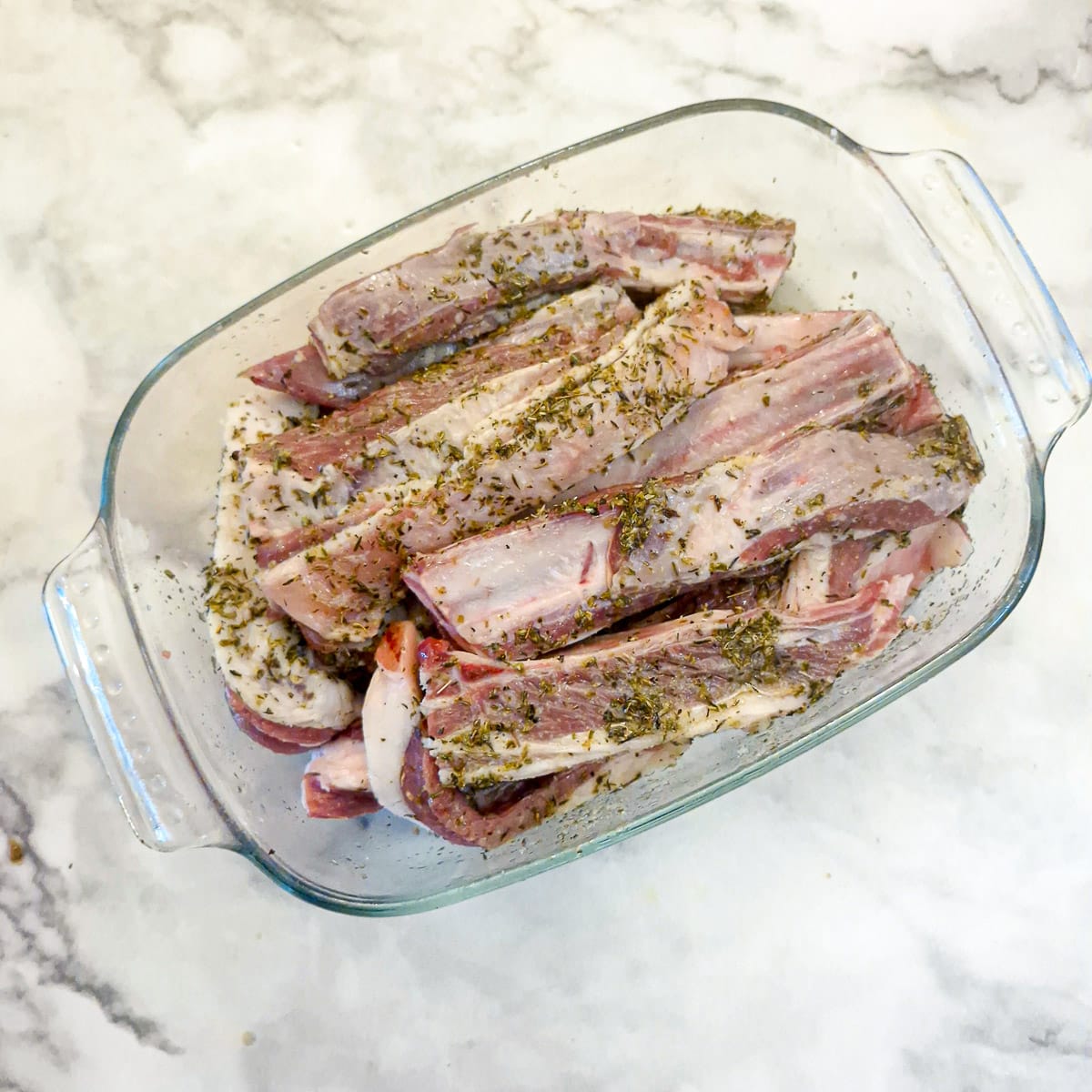 A glass dish of lamb ribs marinating in a lemon and herb marinade.