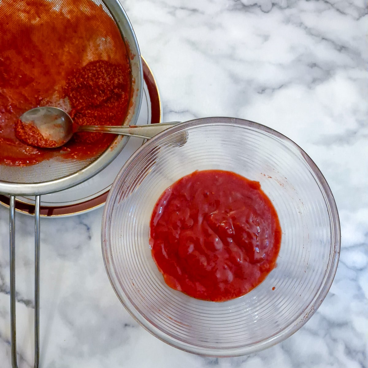 Raspberry pulp in a small glass bowl. There is a sieve in the background containing the seeds that were removed.