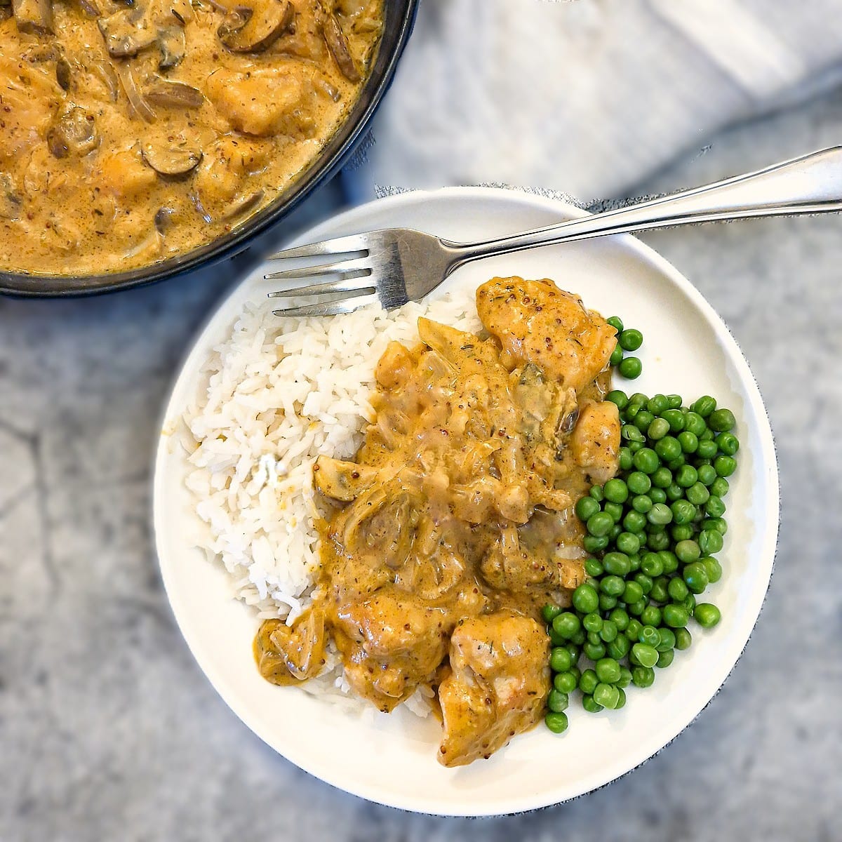 Mushroom chicken with mustard sauce spooned over a helping of rice on a white dinner plate with peas on the side.